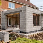 Extension de maison en parpaings en cours de construction contre une habitation ancienne en brique rouge dans le Nord de la France.