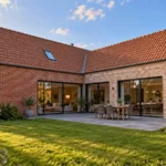 Extension de maison en L dans le Nord, avec façades en briques rouges, toiture en tuiles, grandes baies vitrées et terrasse en pierre bleue donnant sur le jardin.