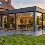 Extension vitrée de plain-pied à toit plat et baies aluminium, accolée à une maison en briques rouges, ouverte sur un jardin